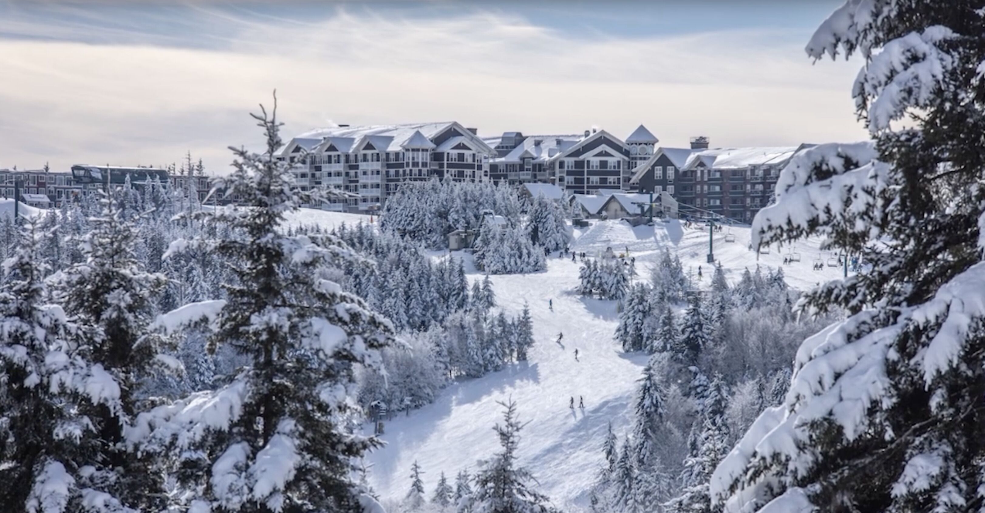 A snow-covered resort with multiple buildings, a ski slope, and dense evergreen trees.