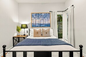 A bedroom with a bed, a painting, a nightstand, a lamp, and a window with curtains.