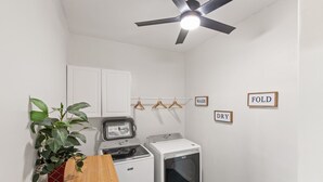 Un lavadero con ventilador de techo, lavadora, secadora y un cesto para ropa que ahorra espacio. Se ven perchas de madera en un colgador y etiquetas que dicen 'LAVAR', 'SECAR' y 'DOBLAR'.