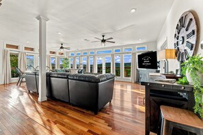 A spacious living room with a large black leather sofa, wooden flooring, and a large wall clock.