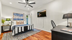 Un dormitorio con cama, escritorio, silla, ventilador de techo, televisor y un cuadro en la pared.