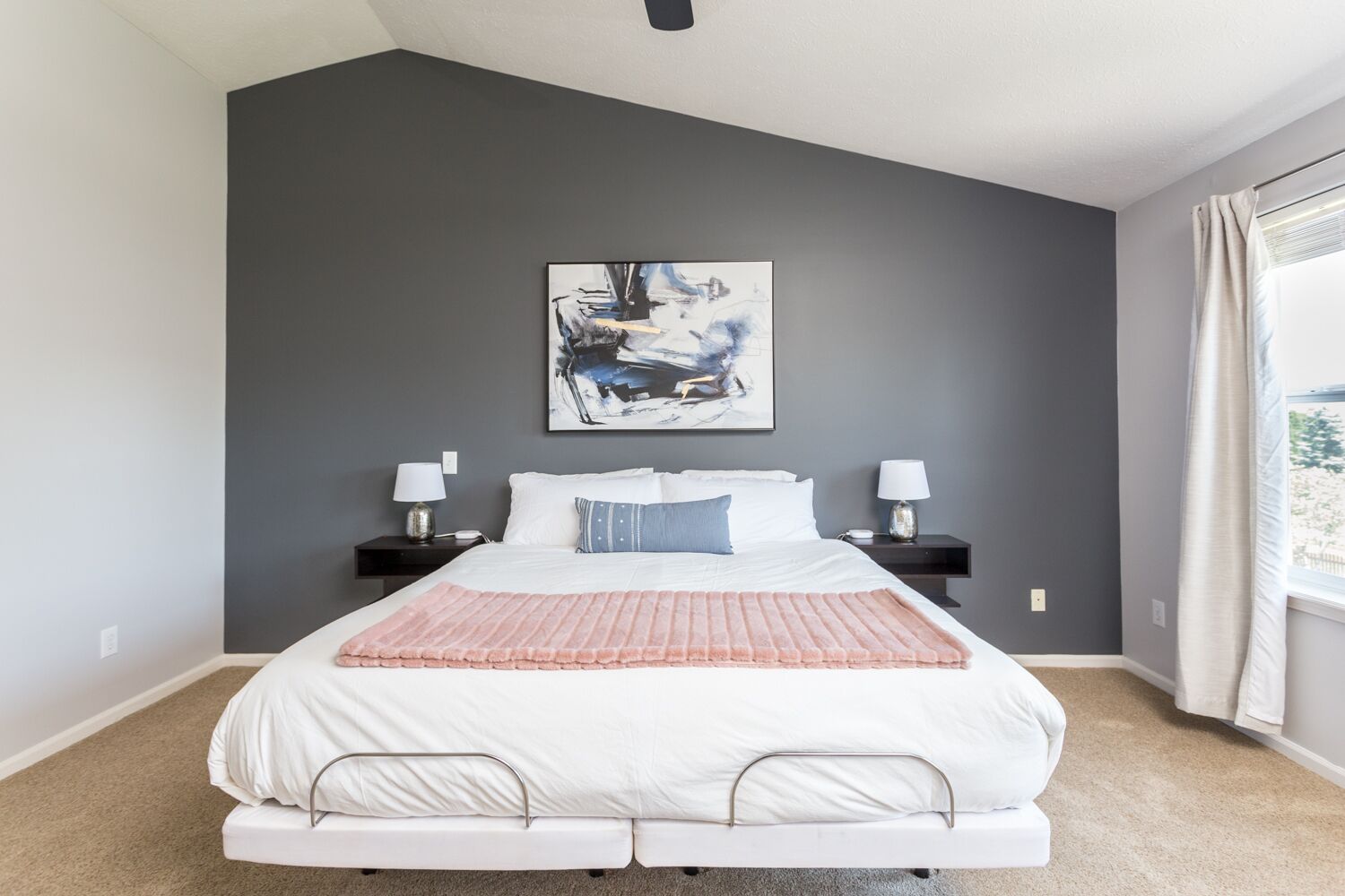 Une chambre moderne avec un grand lit, deux tables de chevet avec des lampes, un mur d’accent de couleur grise et une fenêtre avec des rideaux.