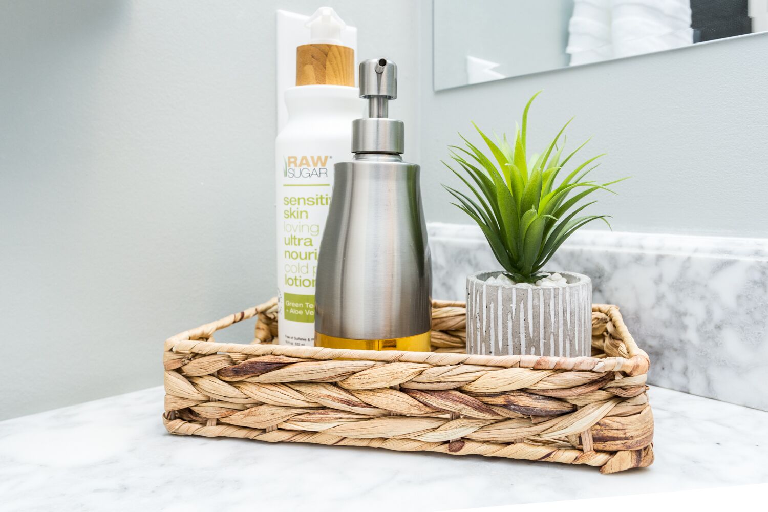 Casa de banho com cesto de fibra natural contendo um frasco de loção, uma pequena planta em vaso e um frasco de produto de cuidados para a pele.