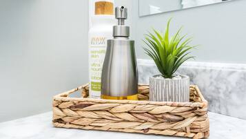 Casa de banho com cesto de fibra natural contendo um frasco de loção, uma pequena planta em vaso e um frasco de produto de cuidados para a pele.