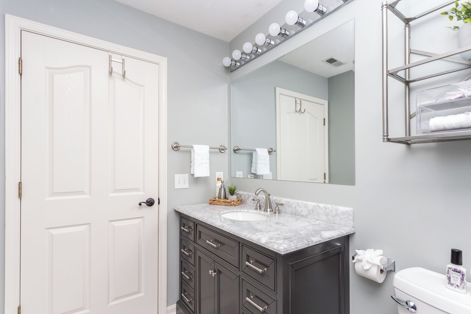 Une salle de bain avec une porte blanche, des meubles sombres, un lavabo en marbre, un grand miroir et une étagère avec des serviettes.