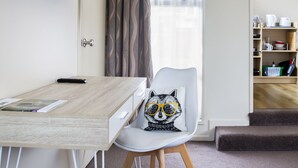 A compact room with a wooden desk, a white chair with a raccoon pillow, a TV mounted above the desk, a window with curtains, and a small kitchenette area. - Chic Private Studio Close to Airport, Bus & Shops - Breakfast included (Christchurch)