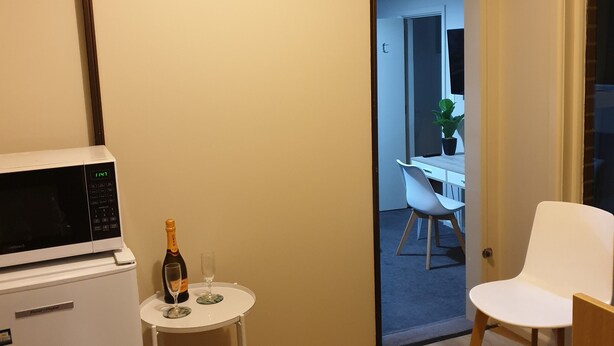 A compact room with a microwave, a small table with a bottle and glass, a white chair, and an air conditioning unit. - Chic Private Studio Close to Airport, Bus & Shops - Breakfast included (Christchurch)