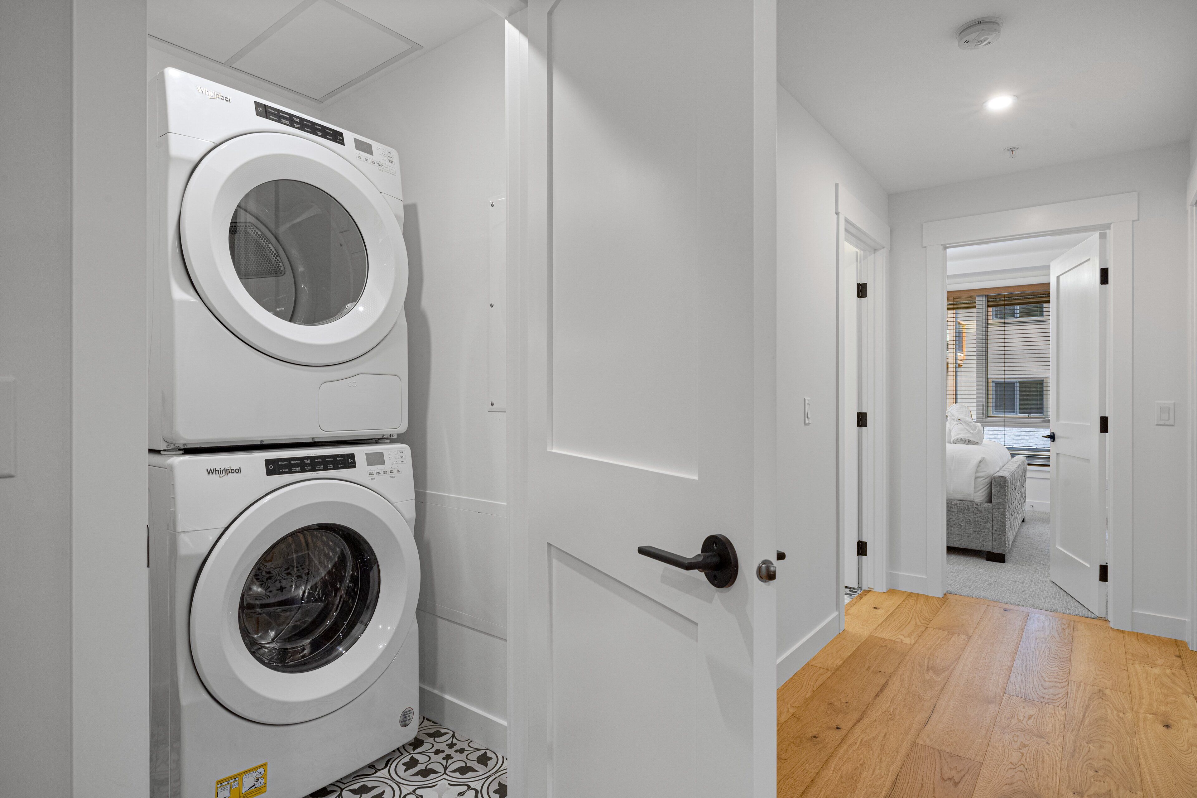 Un lavadero con dos lavadoras y secadoras Whirlpool apiladas, piso de madera y una puerta que da a un dormitorio con cama y ventana.
