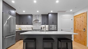 Una cocina moderna con armarios grises, un refrigerador de acero inoxidable, una isla con taburetes y una puerta de madera.