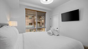 Habitación de hotel con una cama grande, un televisor de pantalla plana montado en la pared, una ventana con persianas y una lámpara en la mesita de noche.