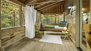 Eine Holzhütte mit Holzvertäfelung, einem Bett mit grüner Bettwäsche, einem kleinen Tisch mit einer Lampe und Blick auf den Wald durch große Fenster.