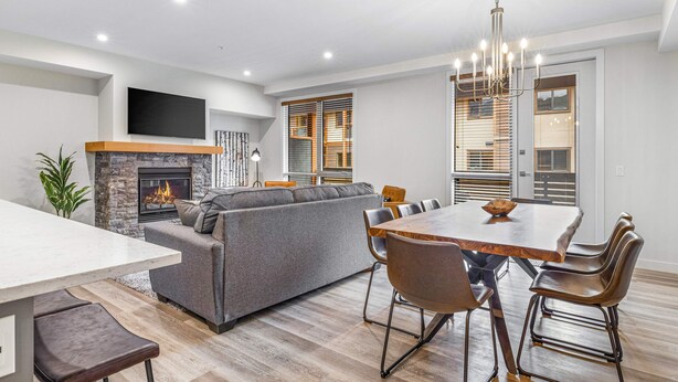 A modern living room with a fireplace, a large gray sofa, a dining table with chairs, and a TV mounted on the wall.