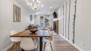 Een moderne eetruimte met een houten tafel en witte stoelen, een kroonluchter en wanddecoratie met afbeelding van berkenbomen.