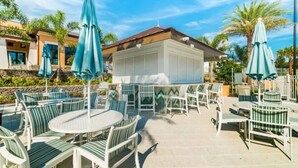 An outdoor patio area with round tables and chairs, surrounded by palm trees and umbrellas. - Solara 7BR/6.5BA Villa w/Pool/Spa, 3544 sqft, Sleeps 18, Lake and green view. (Reunion)