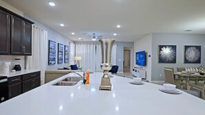 A modern kitchen with a central island, a dining area with a table set for four, and a living room with a TV and artwork. - Solara 7BR/6.5BA Villa w/Pool/Spa, 3544 sqft, Sleeps 18, Lake and green view. (Reunion)