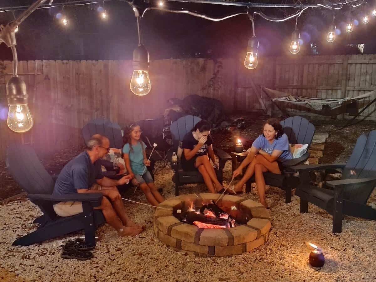 A group of people sitting around a fire pit in an outdoor setting with string lights overhead.