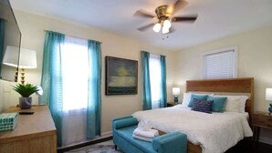 Un dormitorio con cama, escritorio, silla, ventilador de techo y ventana con cortinas.