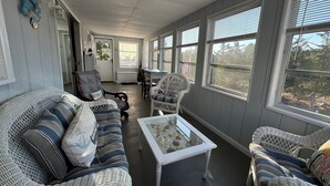 A cozy living room with wicker furniture, a glass coffee table, and large windows offering a view of the outdoors. - Broadkill Beach House- Dog Friendly! Sleeps 13 (Milton)