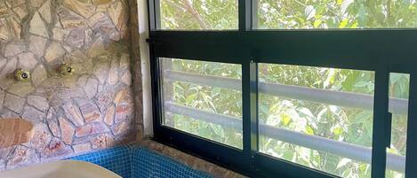 Un baño con pared de piedra, ventana con vista a vegetación, piso de azulejos azules y lavamanos blanco.