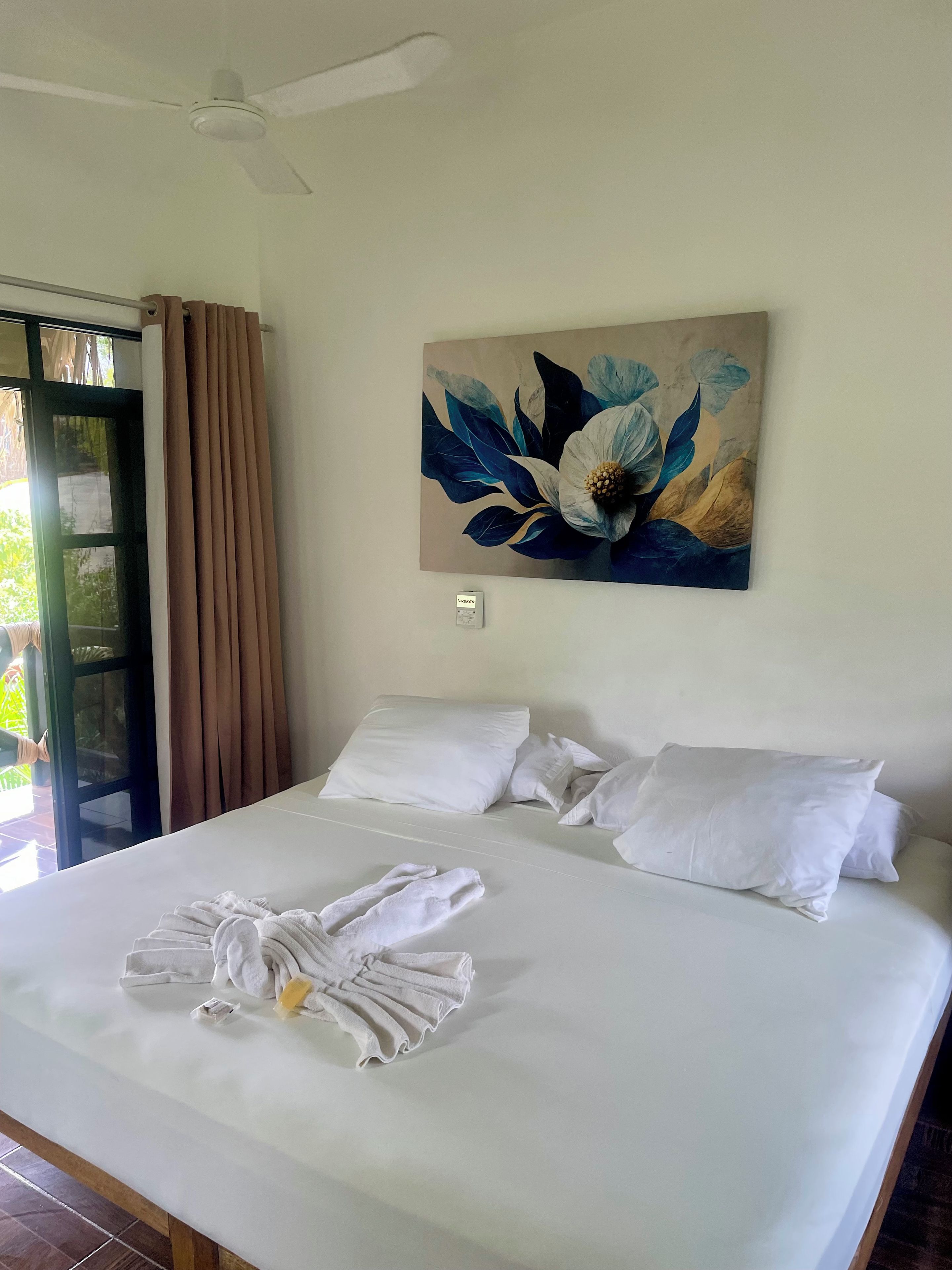 A hotel room with a bed, pillows, a ceiling fan, a window with curtains, and a wall-mounted painting.