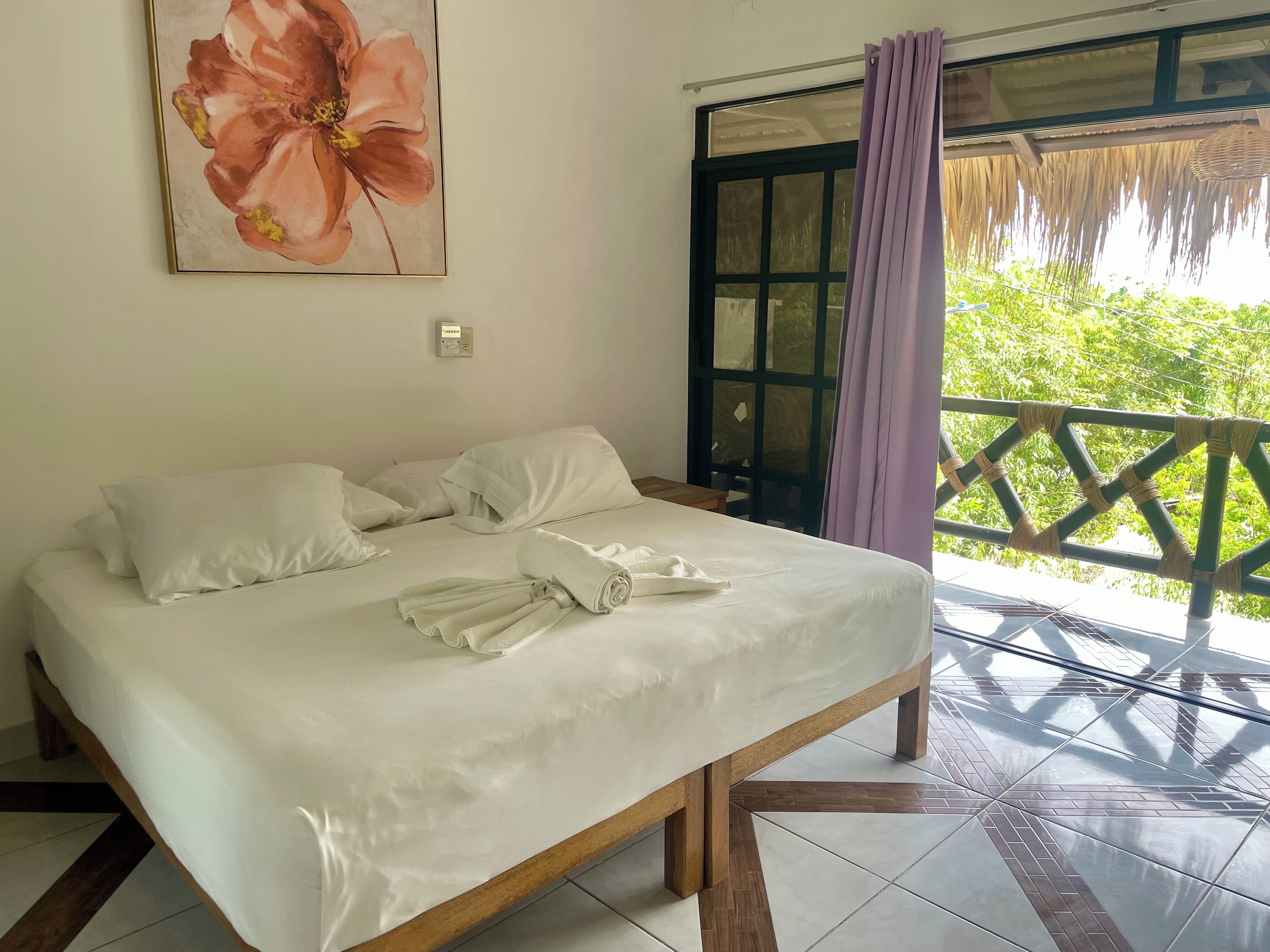 A neatly made bed with pillows and a rolled towel, a painting of a flower on the wall, and a balcony with a view of greenery.