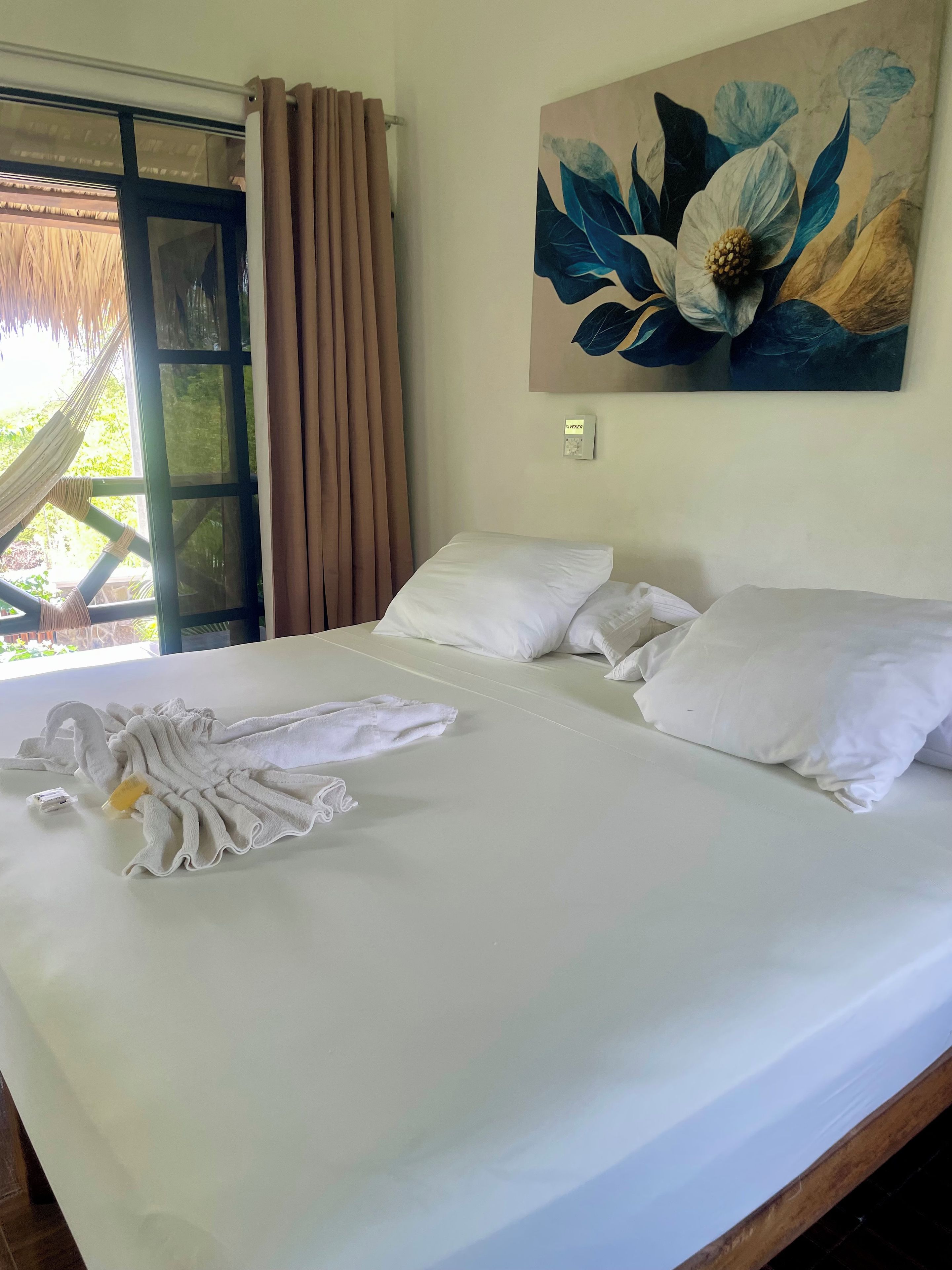 Cama bem arrumada com lençóis brancos e toalha, um quadro de flores na parede e vista para o verde pela janela.