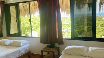 A room with two beds, a ceiling fan, a small table with a radio, and large windows with curtains.