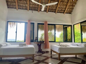 Habitación de hotel con dos camas, ventilador de techo, ventanas grandes con cortinas y vista al mar.