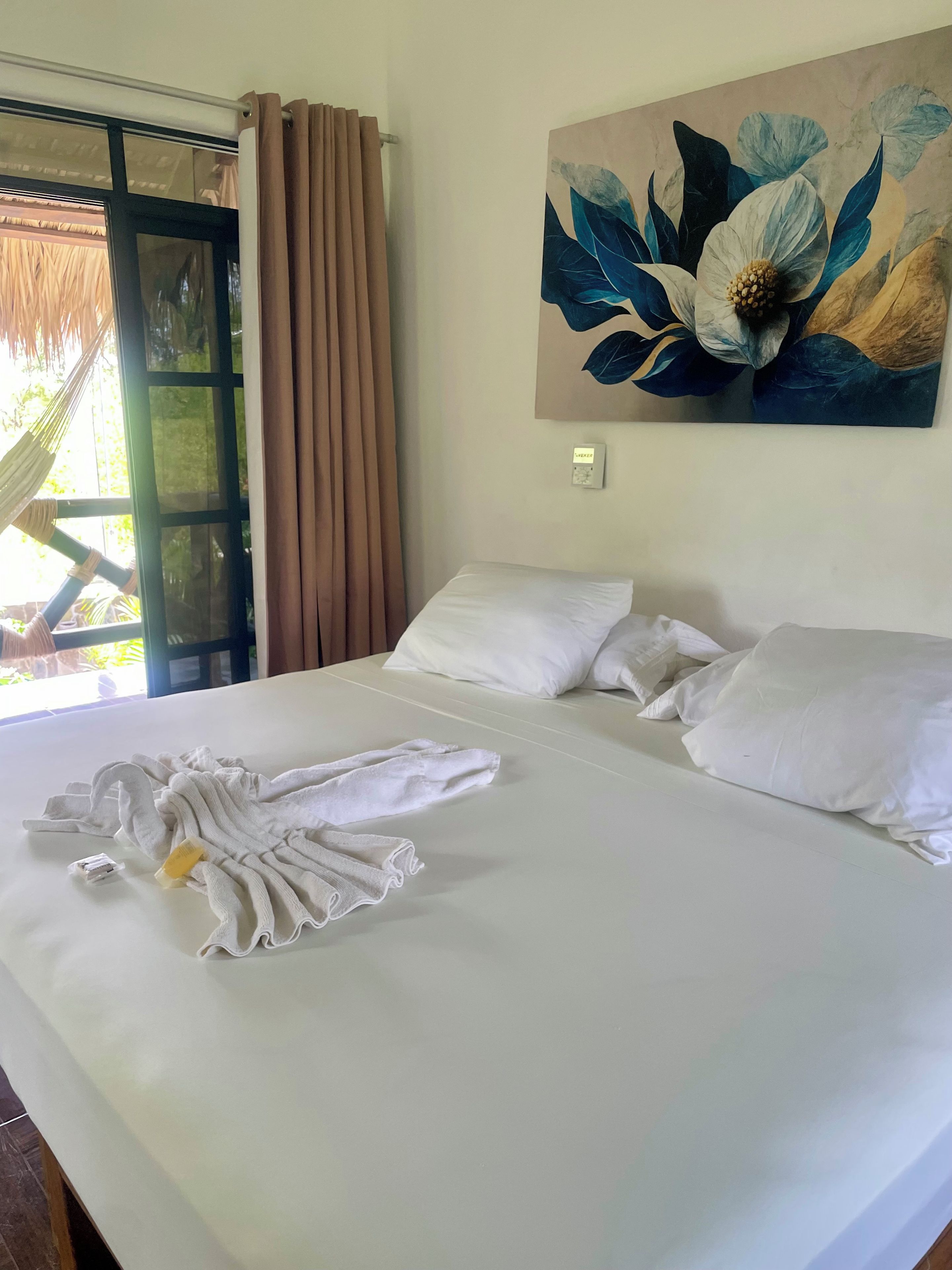 A hotel room with a bed, a painting of flowers, a window with curtains, and a hammock outside.
