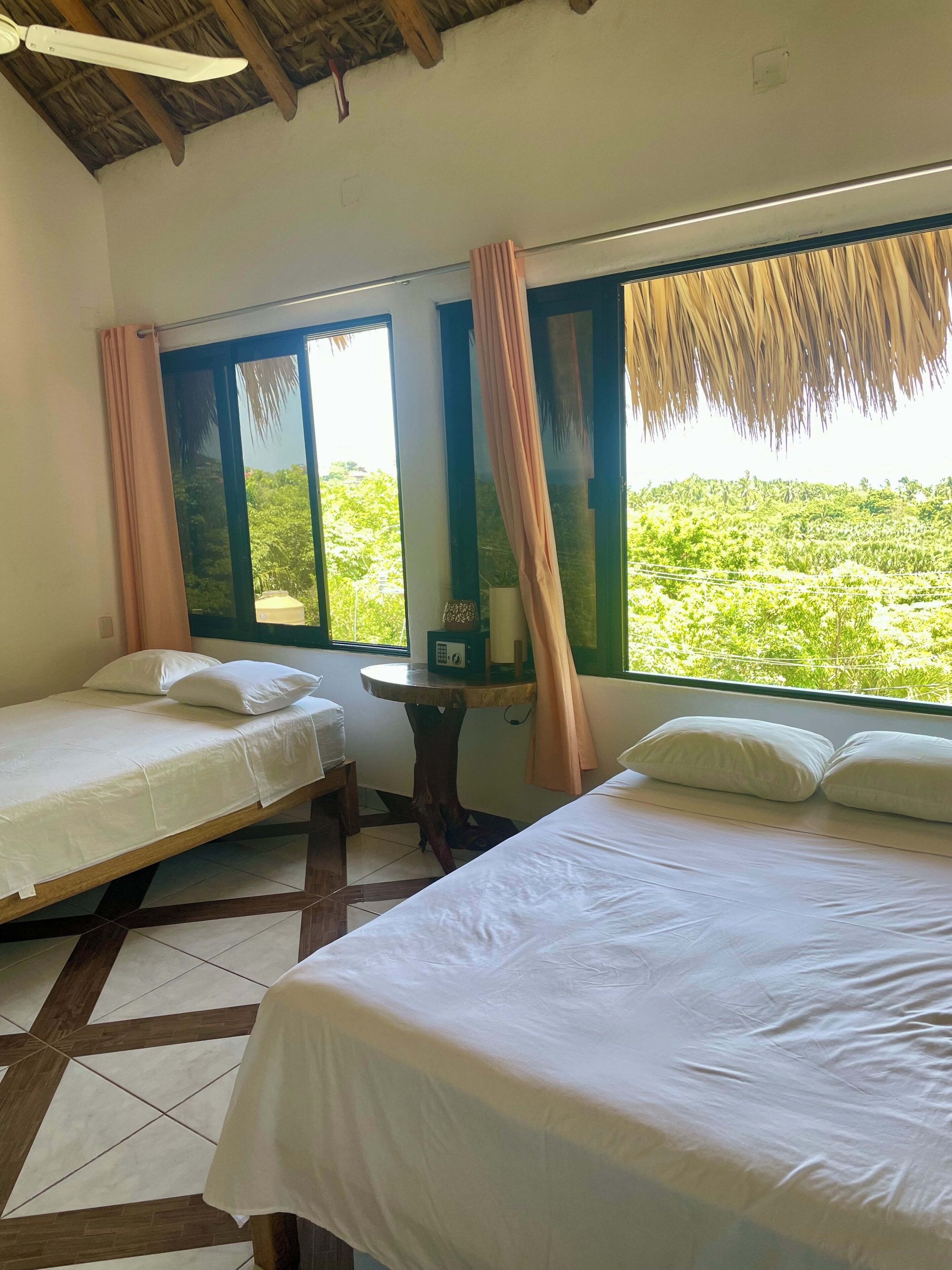Una habitación con dos camas, una mesa y un ventanal con cortinas.