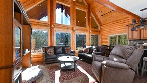 A spacious log cabin interior with a high ceiling, large windows, a cozy seating area with leather sofas, and a kitchen area. - Log Cabin Getaway: WIFI, GameRoom, Firepit, pets OK! (Cascade)