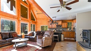 A spacious living room with a fireplace, a ceiling fan, and a kitchen area with wooden cabinets and a stove. - Log Cabin Getaway: WIFI, GameRoom, Firepit, pets OK! (Cascade)