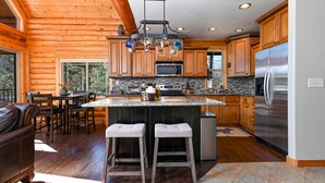A spacious kitchen with wooden cabinets, a large island with stools, a stainless steel refrigerator, and a stone backsplash. - Log Cabin Getaway: WIFI, GameRoom, Firepit, pets OK! (Cascade)