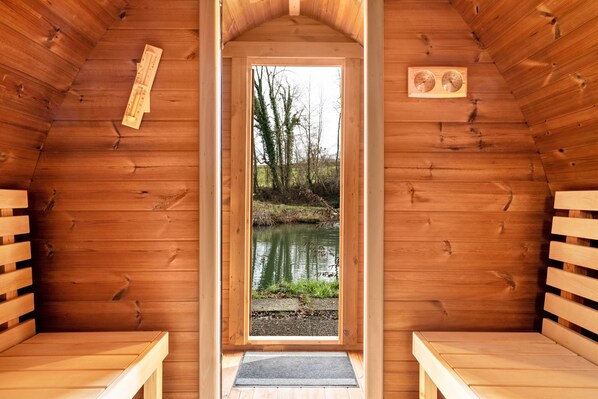 A wooden sauna with benches, a door leading to a natural outdoor area with a pond, and a wooden sign on the wall.