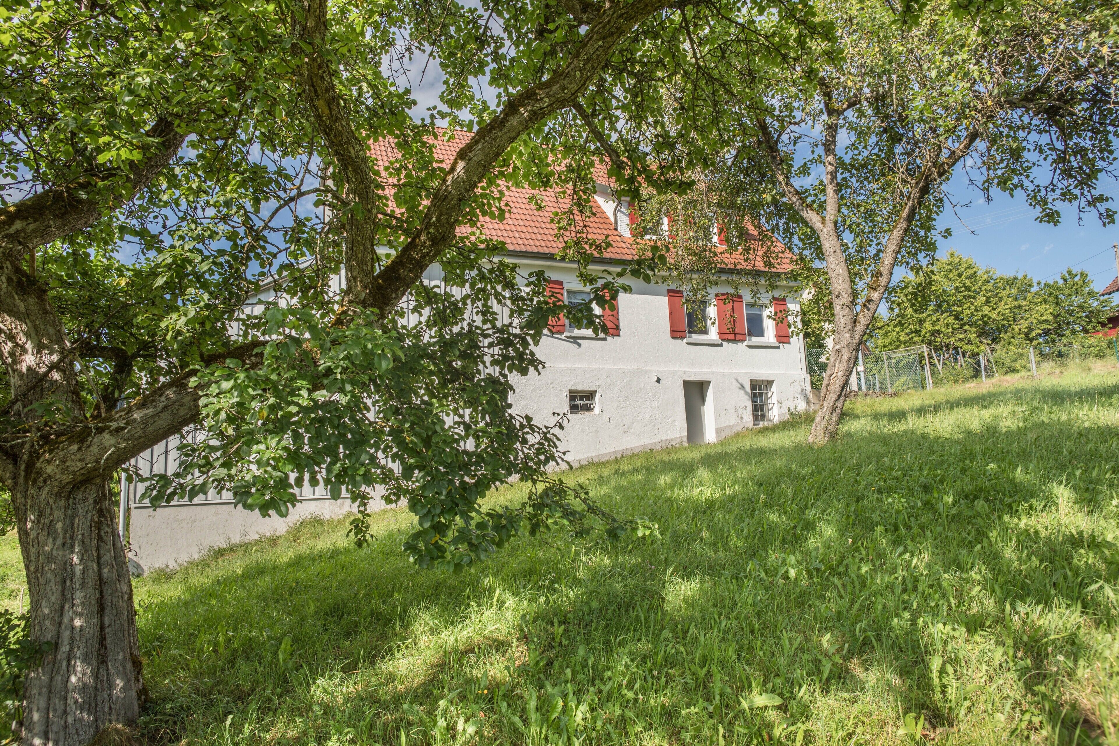 Une maison avec un toit recouvert de tuiles rouges, entourée d’une végétation luxuriante et d’arbres.