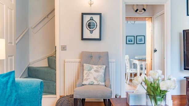 A living room with a grey armchair, a blue sofa, and a wooden coffee table. There is a framed picture on the wall and a vase with white flowers on the table.