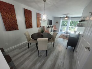 Uma sala de jantar com uma mesa redonda, quatro cadeiras e um candelabro. Dá para ver uma cozinha com geladeira e mesa de jantar ao fundo.