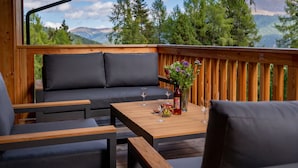 A wooden deck with a sofa and chairs, a table with wine glasses and a vase of flowers, and a view of trees and mountains.