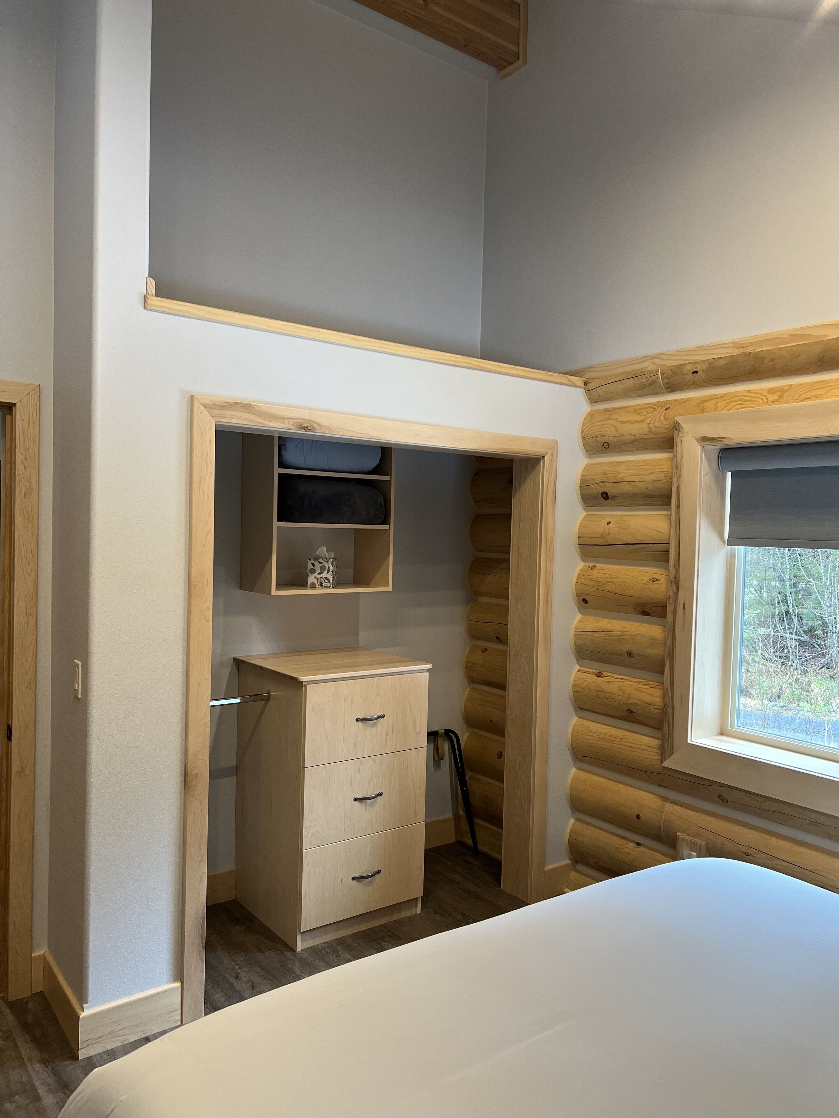 A bedroom with a bed, a wooden dresser, a shelf with cushions, and a window with a view of trees.