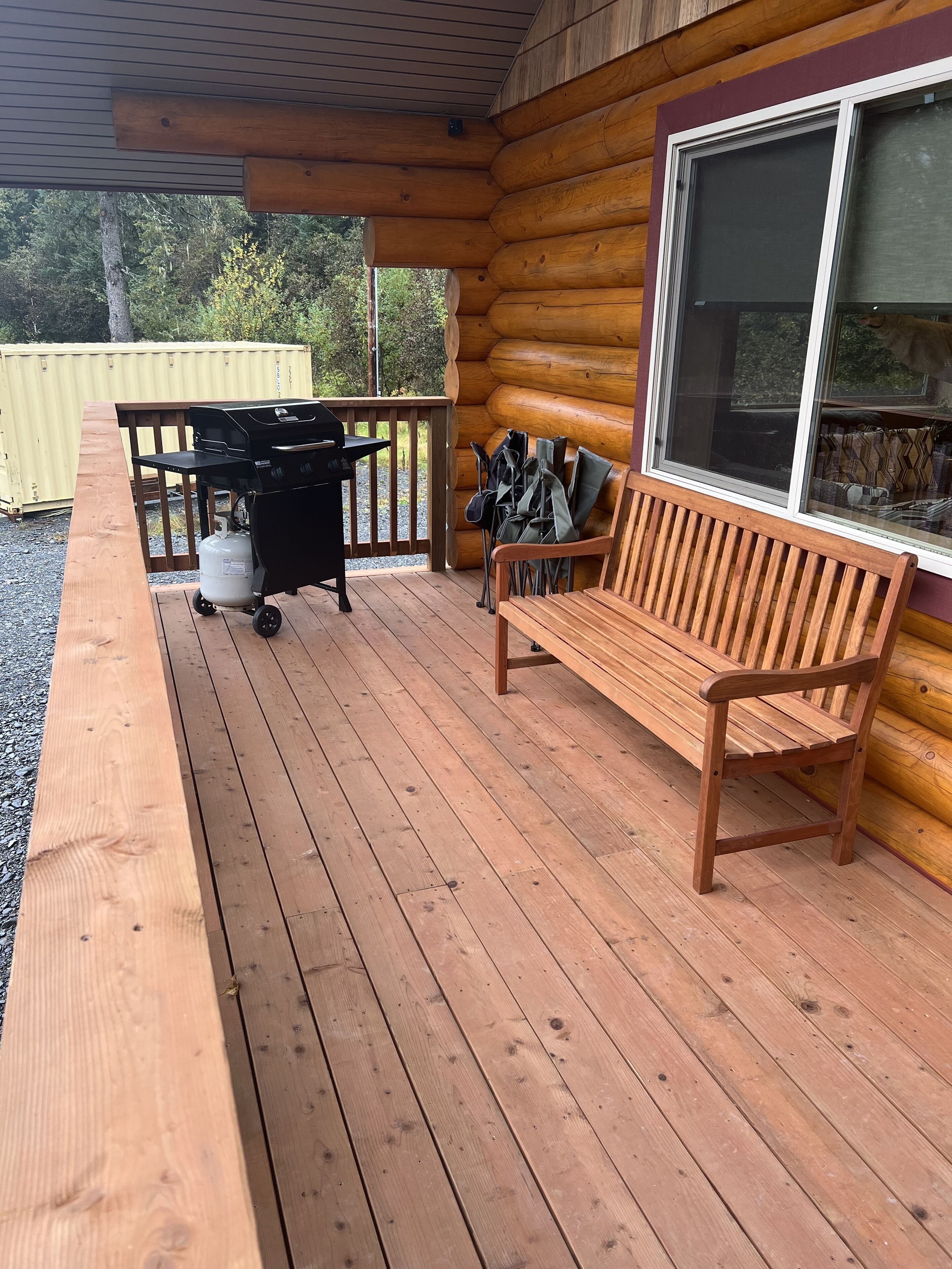 A wooden deck with a grill and benches, a large window, and a view of trees and a shipping container in the background.