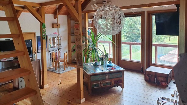 A spacious room with wooden beams, a loft area, a hanging decorative light, a wooden table with plants, and a large window with a view. - House in Teichland Pool, sauna, fireplace on the Cottbus Baltic Sea with proximity to the Spreewald (Teichland)