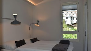 Une chambre moderne avec un lit, deux lampes fixées au mur et une fenêtre donnant sur des bâtiments et de la verdure.
