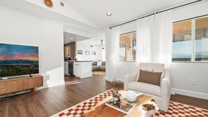 Un salon moderne doté d’un grand téléviseur à écran plat, d’un canapé blanc, d’une table basse avec un magazine, et offrant une vue sur la cuisine et l’espace repas grâce à une grande fenêtre.