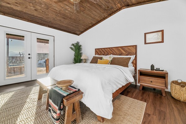 A bedroom with a wooden ceiling, a bed with white bedding, a wooden nightstand, a small table with a bowl, and a woven basket.