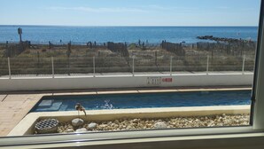 Ein Pool mit einem Vogel am Rand, Blick auf das Meer und ein Zaun, der den Poolbereich vom Strand trennt.
