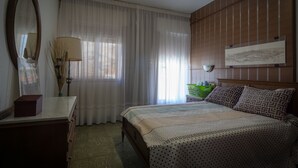Ein Schlafzimmer mit einem Bett, einer Kommode, einem Spiegel, einem Fenster mit durchsichtigen Vorhängen und einem an der Wand hängenden Kunstwerk.