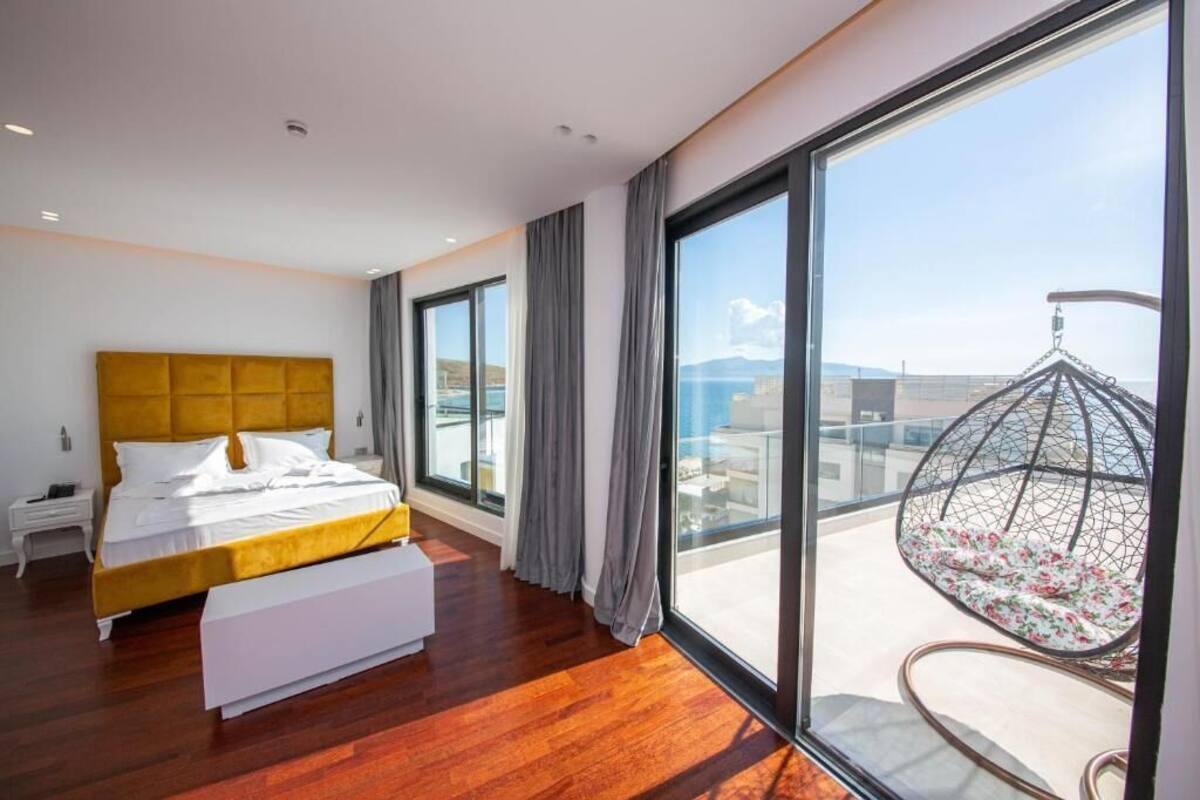 A modern bedroom with a large bed, a white bedside table, a bench, and a view of the city through large windows.