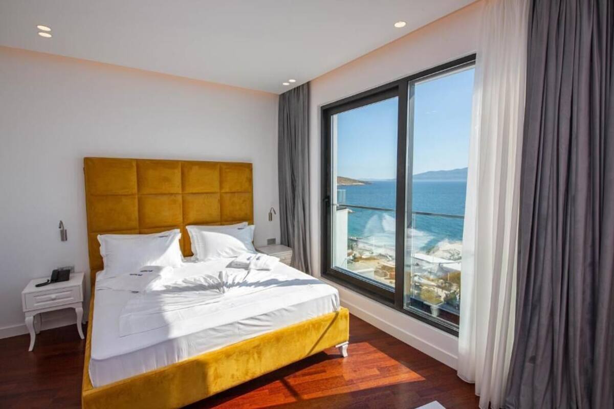 A modern hotel room with a large bed, a nightstand, and a view of the sea through a large window.