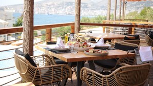 Un comedor con sillas y mesas de mimbre, vista al mar y montañas al fondo.