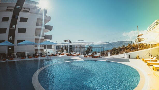 Una piscina con zonas de sol y sombrillas, rodeada de edificios con balcones y vistas al mar.